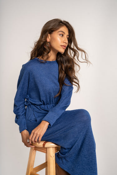 Woman wearing a blue dress sitting on a wooden stool against a plain background