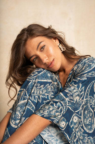 A woman with wavy brown hair wearing a blue and white paisley patterned top, leaning her head on her hand against a textured beige background.