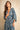 A woman wearing a blue and white paisley print kaftan dress with wide three-quarter sleeves, a deep V-neckline, and a gathered waist, posed against a textured beige background.