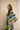 A woman wearing a loose, patterned kaftan dress with a geometric design in yellow, navy, and light blue, standing against a textured beige wall.
