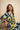A woman wearing a colorful abstract print caftan dress sits on a wooden chair against a textured beige wall.