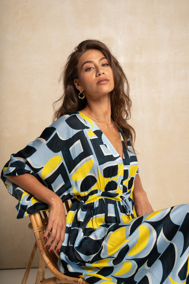 A woman wearing a colorful abstract print caftan dress sits on a wooden chair against a textured beige wall.