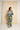 A woman stands with her back to the camera wearing a matching two-piece lounge set with a bold abstract geometric pattern in yellow, navy, and light blue. She is positioned in front of a large, textured beige canvas against a white studio wall.