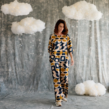 Woman in a patterned top and pants standing in front of a textured wall with cloud decorations.