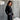 Woman in a black outfit standing against a textured gray wall with white clouds.
