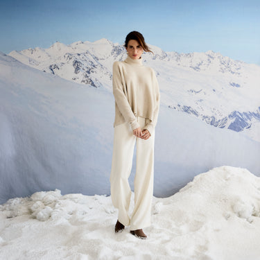 Person wearing a beige sweater and white pants standing on a snowy mountain