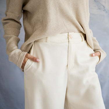 Person wearing a beige sweater and cream pants against a gray background