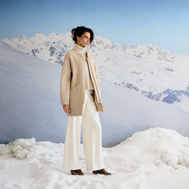 Person wearing a beige coat and white pants standing in a snowy landscape with mountains in the background