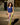 A woman stands on a city street wearing a sleeveless royal blue top and high-waisted, multicolored striped pants with a brown belt. The background features a patterned wall and a window with gold-toned circular details.