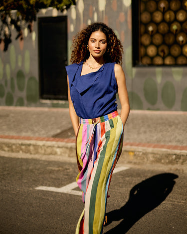 A woman stands on a city street wearing a sleeveless royal blue top and high-waisted, multicolored striped pants with a brown belt. The background features a patterned wall and a window with gold-toned circular details.