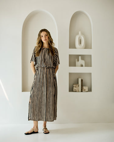 A woman modeling a short-sleeve midi dress with vertical black and beige stripes, standing in a minimalist white room with arched recessed shelves.