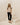 A woman stands on white geometric steps wearing a beige short-sleeve top with black shoulder panels, black cropped pants, and black sandals in a minimalist cream-colored room.