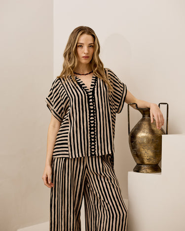 A woman models a matching two-piece set featuring a black and beige striped short-sleeve top and wide-leg pants, posed next to a decorative bronze vase on a white pedestal.