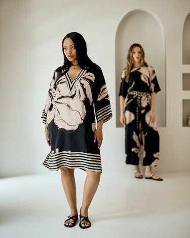 A woman models a loose black tunic dress with a beige floral print and striped hem in a bright studio with arched alcoves in the background. Another woman stands blurred in the back wearing a matching set.