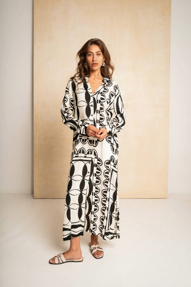 A woman modeling a long-sleeved maxi shirt dress with a bold black and white abstract geometric pattern, standing against a minimalist beige backdrop.