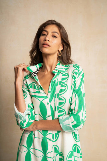 A woman wearing a green and white patterned button-up shirt dress, posing against a textured beige background.