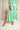 A woman wearing a long-sleeve maxi dress with a bold green and white geometric pattern, paired with metallic gold sandals, standing in front of a neutral beige backdrop.