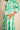 A close-up of a person wearing a long-sleeved shirt dress with a bold green and white geometric floral pattern, featuring a side pocket with the hand resting inside.