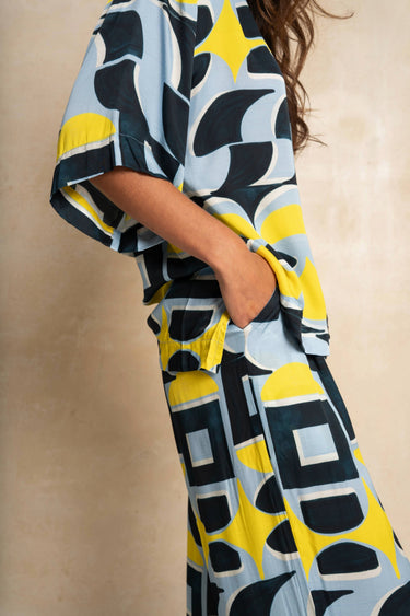 A woman wearing a loose, short-sleeve top and matching pants with a bold abstract geometric print in yellow, navy blue, and light blue, standing against a neutral beige textured wall.