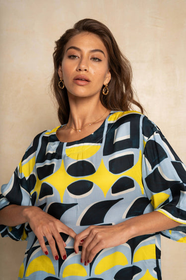 A woman wearing a loose-fitting short-sleeve top with a bold abstract geometric pattern in light blue, yellow, and navy, posed against a neutral beige textured background.