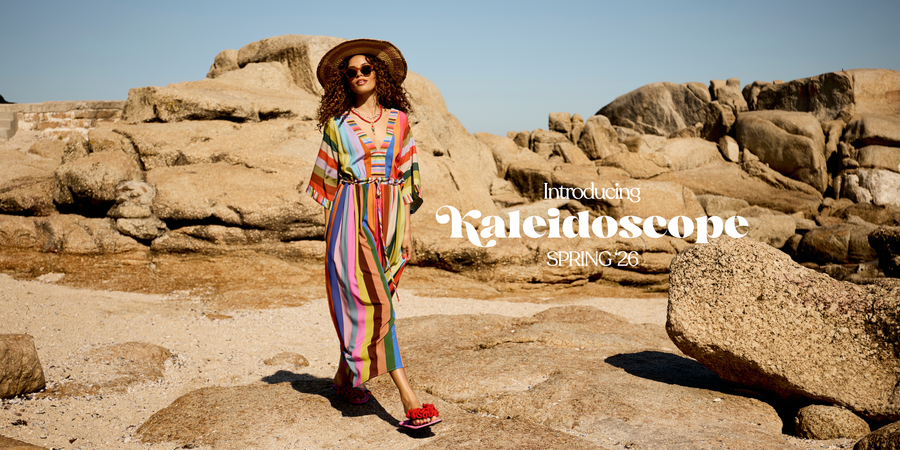 A woman wearing a colorful striped bohemian jumpsuit, wide-brimmed straw hat, and sunglasses stands on a rocky beach with large tan boulders under a clear blue sky. White text on the right reads 'Launching Kaleidoscope SPRING 26'.