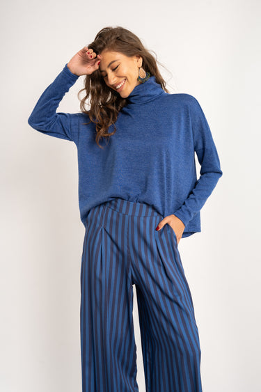 Woman wearing a blue cowl-neck sweater and striped pants against a white background