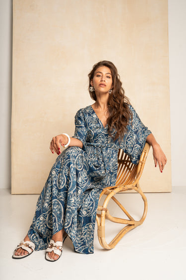 A woman wearing a blue and white paisley print kaftan dress sits on a wooden chair against a beige textured backdrop.