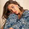 A woman with wavy brown hair wearing a blue and white paisley patterned top, leaning her head on her hand against a textured beige background.