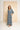 A woman modeling a blue and white paisley print maxi dress with wide sleeves and side pockets, standing in front of a large beige backdrop panel.