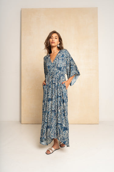 A woman modeling a blue and white paisley print maxi dress with wide sleeves and side pockets, standing in front of a large beige backdrop panel.