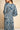 A woman shown from the back wearing a long-sleeved maxi dress with a blue and white paisley block print pattern, set against a neutral beige background.