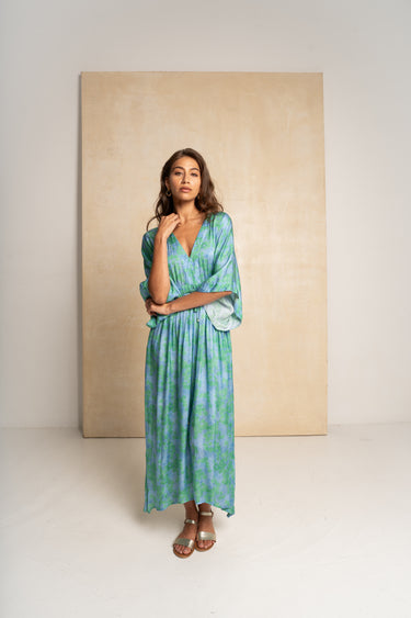 A woman modeling a long, flowing maxi dress with a green and blue tie-dye pattern, standing in front of a large beige canvas print on a white studio background.