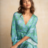 A woman wearing a flowing kaftan dress with a green and blue tie-dye pattern, posed against a textured beige background.