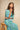 A woman wearing a flowing green and blue patterned kaftan dress sits on a wooden stool against a textured beige wall.