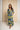 A woman modeling a long-sleeve maxi dress with a bold geometric pattern in yellow, black, and light blue-gray, standing against a textured beige backdrop.
