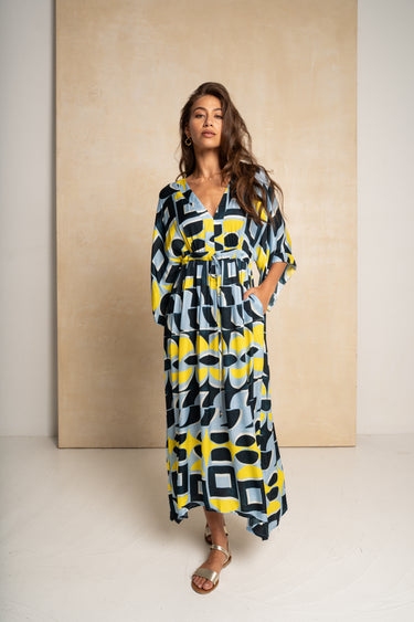 A woman modeling a long-sleeve maxi dress with a bold geometric pattern in yellow, black, and light blue-gray, standing against a textured beige backdrop.