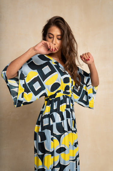 A woman wearing a loose dress with a bold abstract geometric pattern in yellow, black, and light blue-gray, posed against a warm beige textured backdrop.