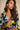 A woman wearing a loose-fitting V-neck top with a bold abstract geometric pattern in navy blue, light blue, yellow, and white, posed against a neutral beige background.