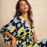 A woman wearing a colorful abstract print caftan dress sits on a wooden chair against a textured beige wall.