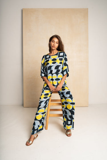 A woman wearing a matching two-piece set with a bold abstract geometric pattern in yellow, navy, and light blue, sitting on a wooden stool in front of a beige backdrop.