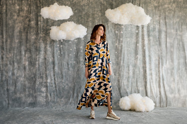 Woman in a patterned dress standing in front of a textured wall with cloud decorations.
