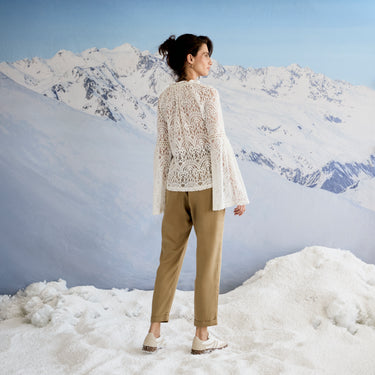 Woman wearing a white lace cardigan and beige pants standing in a snowy landscape with mountains in the background.