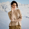 Woman wearing a beige sweater and pants standing in front of snow-covered mountains.