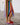 A person wearing a long, flowing maxi dress with vertical rainbow stripes in pink, yellow, green, blue, orange, and brown, paired with bright pink flip-flops featuring red tassels, standing on a stone paved surface in front of a white garage door.