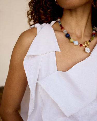 A close-up of a model wearing a white draped top and a colorful beaded necklace with gold-toned accents and a white shell pendant.