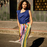 A woman stands on a city street wearing a sleeveless royal blue top and high-waisted, multicolored striped pants with a brown belt. The background features a patterned wall and a window with gold-toned circular details.