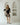 A woman models a black and beige abstract floral print tunic dress with striped details, standing in a minimalist white room with arched recessed shelves.