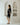 A woman stands in profile wearing a black and beige abstract-patterned tunic dress with striped hem and sleeves, paired with black sandals, set against a minimalist white wall with arched recesses.