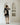 A woman stands with her back to the camera wearing a black short-sleeve tunic dress with large beige floral motifs and striped trim, set against a minimalist white wall with built-in arched shelves.