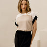 A woman modeling a color-blocked short-sleeve blouse with a white and beige yoke and black sleeve panels, paired with black trousers, in a minimalist cream-colored room with geometric steps.